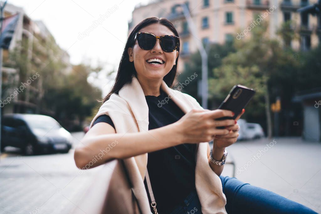 Beautiful trendy dressed female blogger laughing on funny content using mobile phone on free time outdoors, smiling woman 20s enjoying 4G connection on street in roaming chatting via application
