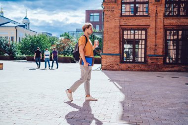Gündelik giyinmiş, gözlüklü, ders kitabı ve sırt çantalı, parlak güneşin altında kahve içen sakin bir adamın yan görüntüsü.