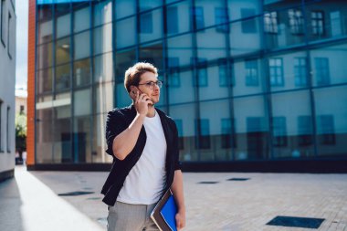 Yoğunlaşmış, gözlüklü, koyu renk ceketli ve beyaz tişörtlü bir erkek. Gündüz vakti elinde defterlerle sokakta dikilip gözlerini kaçırırken cep telefonundan arıyor.