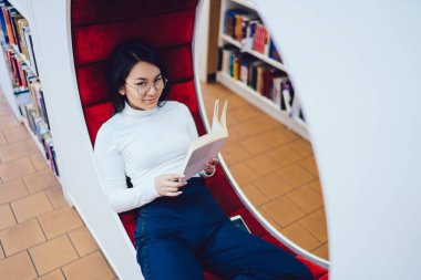 Modern kütüphanede moda bankta dinlenen ve okunabilir kitapların keyfini çıkarırken kameraya bakan gözlüklü zeki etnik kadının yüksek açısı.