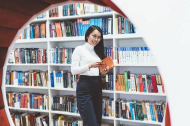 Sıradan kıyafetleri ve gözlükleriyle gülümseyen Asyalı kadın kitapçıda elinde kitaplıkla kameraya bakarak kitabı tutuyor.