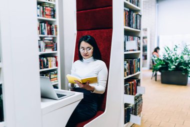 Kitapta bilgi arayan düşünceli Asyalı kadın ve dizüstü bilgisayarla kütüphanede kırmızı yumuşak sırtı olan bir sandalyede çalışıyor.