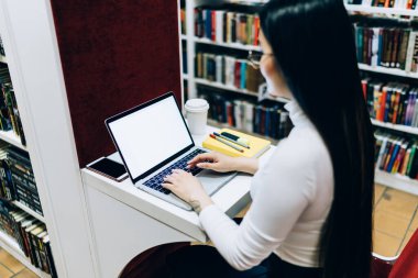 Tek tek kütüphane masasında otururken ders yazarken boş ekranla bilgisayarını karıştıran siyah saçlı kız öğrencinin yan görüntüsü