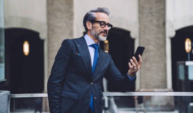 Gri ofis binasının karşısındaki şeffaf balkonda cep telefonu kullanan şık takım elbiseli ve gözlüklü ciddi bir orta yaşlı işadamı.