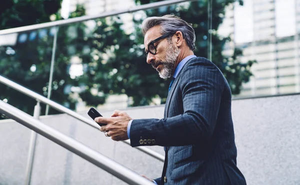 从下面的侧面看英俊的中年男性 身穿正式西装 戴着眼镜 走上楼去 在城市背景下使用现代数码智能手机 — 图库照片