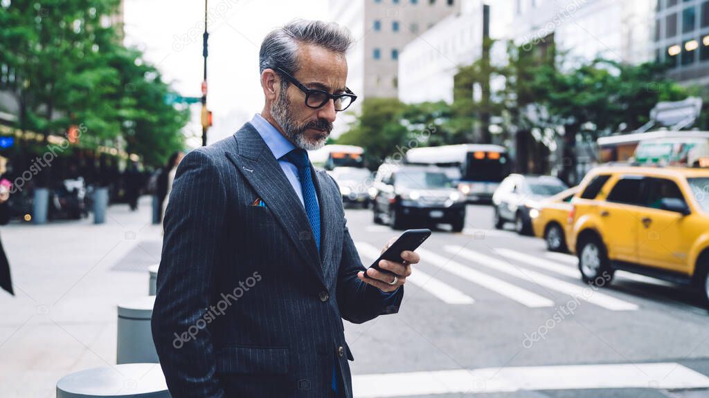 Middle aged businessman in glasses and stylish dark striped business suit standing in busy city street and browsing smartphone in New York