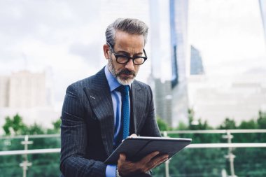 Odaklanmış orta yaşlı yakışıklı erkek resmi temiz takım elbiseli internette geziniyor ve New York 'taki kentsel arka plan hakkında modern dijital cihazlarda bilgi topluyor.