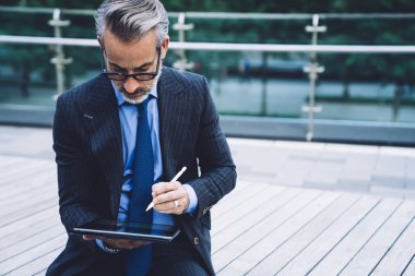 Gözlüklü ve takım elbiseli orta yaşlı bir adam New York 'ta köprü bankında otururken stilus kalemle tablet kullanıyor.