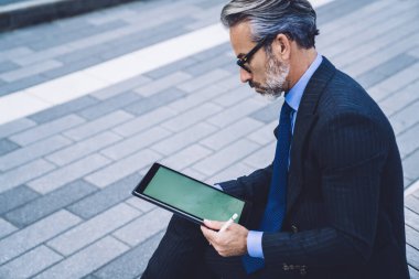 New York caddesindeki bankta otururken, tabletle çizimler üzerinde çalışarak bacak bacak bacak üstüne atmış, konsantre olmuş orta yaşlı işadamının yan görüntüsü