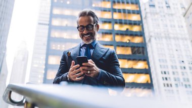 New York gökdelenlerinin arka planında merdiven tipi tırabzanın yanında duran olgun bir erkeğin yanından akıllı telefon ekranına neşeyle bakıyor.