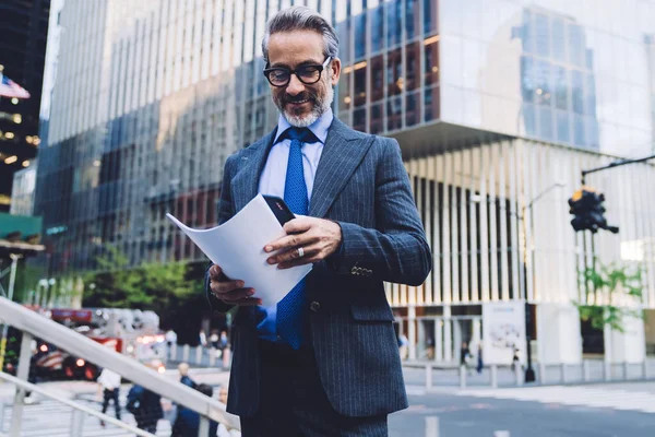 Smiling middle aged man in elegant suit and glasses with documents and mobile phone standing among glass skyscrapers in New York