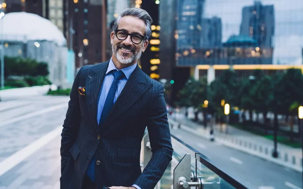 Handsome bearded middle aged man in glasses and business suit standing against evening New York street while looking at camera