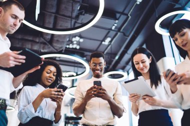 Resmî kıyafetler içinde gülümseyen çok kültürlü insanların düşük açısı. Tabletleri ve akıllı telefonları karıştırırken modern şehir ofisinde bir arada duruyorlar.