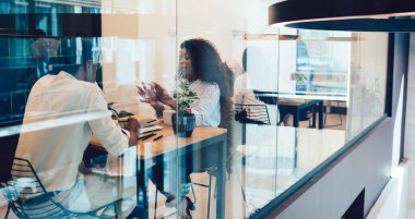 Çok ırklı çalışanlar resmi kıyafetler giyip ellerinde belgeler ve cihazlarla masada oturuyorlar. Kahve içip sohbet ediyorlar.