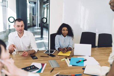 Profesyonel ve prestijli çok ırklı iş arkadaşları ellerinde cihazlar ve çizelgelerle masanın önünde oturmuş yeni iş projesini tartışıyorlar.