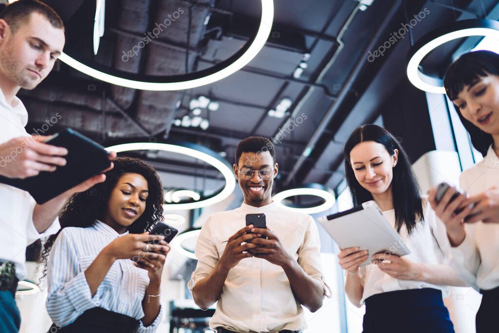 Low angle of positive smiling multiracial people in formal clothes browsing tablets and smartphones while standing together in circle in modern urban office