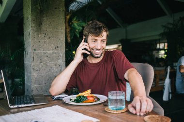 Neşeli Kafkas erkek serbest çalışan cep telefonuyla konuşuyor, kafenin içinde yemek yiyor, pozitif gülümseyen hipster adam akıllı telefondan keyif alıyor ve boş zamanlarında internetten sohbet ediyor.