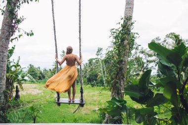 Çağdaş kadın, sallanan portakal rengi yaz kıyafetiyle Palm Grove 'da salıncakta durup tatilin tadını çıkarıyor. 