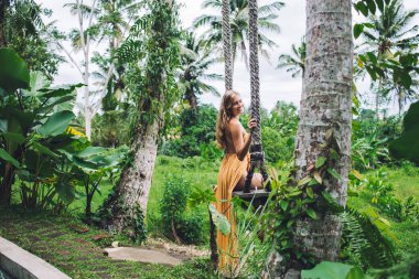 Turuncu yaz kıyafetli neşeli sarışın kıvırcık kadın tropik bahçede çimenlerde salıncakta sallanıyor ve kameraya bakarak tatilin tadını çıkarıyor.