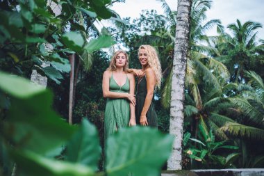 Sarı kıvırcık saçlı, zarif siyah elbiseli yeşil elbiseli, tropik egzotik ormanın yanında elini omzuna koyup kameraya bakan neşeli bir kadın.