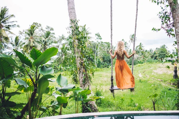 Turuncu yazlık elbiseli rahat sarışın bir kadın tropikal yeşil bahçede salıncakta sallanıyor ve tatil yapıyor.