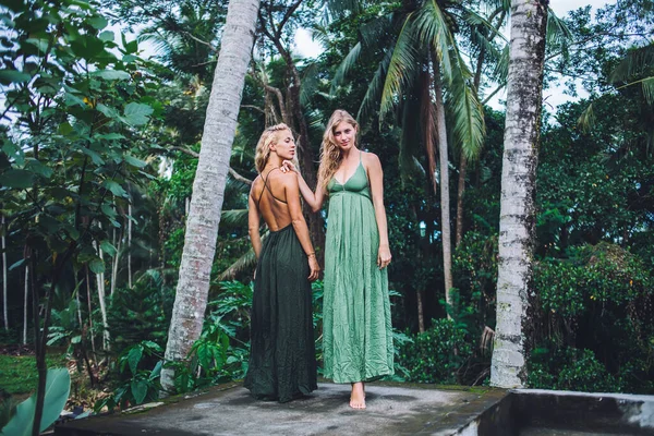 若いです流行の女性見ますカメラと置く手上の肩巻き毛の女性横に立って光同じ夏のドレスで熱帯の背景 — ストック写真