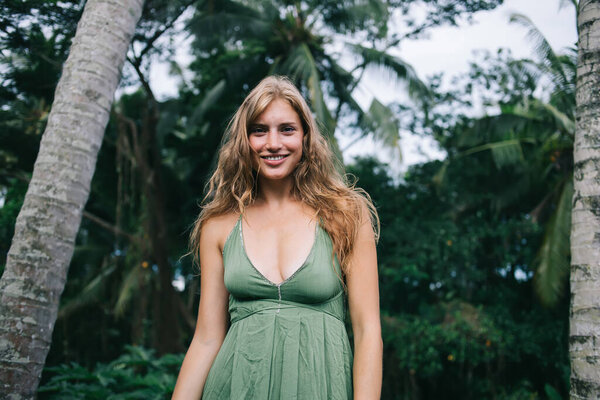 Красивая улыбающаяся молодая женщина с длинными светлыми волосами и натуральным макияжем в зеленом платье с декольте на фоне зеленых деревьев