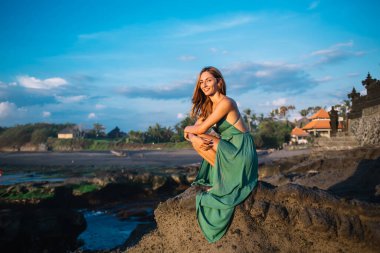 Yeşil elbiseli muhteşem bir kadın vücudu kayalık sahilde oturuyor ve gülümseyip kameraya bakarken güneşli havanın tadını çıkarıyor.