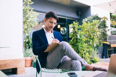 Başarılı modaya uygun giyinmiş modaya uygun erkek yazar dizüstü bilgisayarla terasta yazı hazırlıyor, kendine güvenen işadamı teknoloji üzerinde çalışırken planlarını kontrol ediyor.