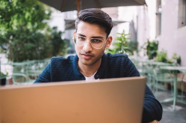 Kafe terasında çalışmak ve kameraya bakmak için dizüstü bilgisayar kullanan rahat giyinen ve gözlüklü kendinden emin genç bir adam.