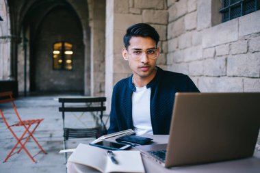 Günlük kıyafet ve gözlüklü genç adam yaratıcı çalışma alanı projesinde çalışırken dizüstü bilgisayar ve günlükle masada oturuyor.