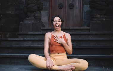 Aktif giyinmiş beyaz kadın portresi boş zamanın keyfini çıkarıyor ve bir şeye şaşırıyor. Komik, genç bir kadın Lotus 'ta oturmuş gülümsüyor ve kameraya bakıyor.