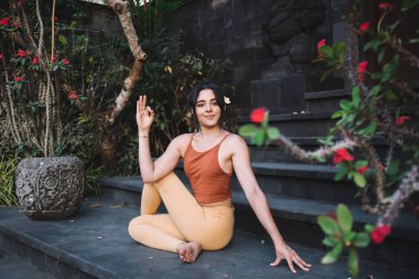 Rahat giyinen genç etnik kadın Ardha Matsyendrasana Poz verirken Bali tatil köyündeki egzotik binaların merdivenlerinde yoga yaparken