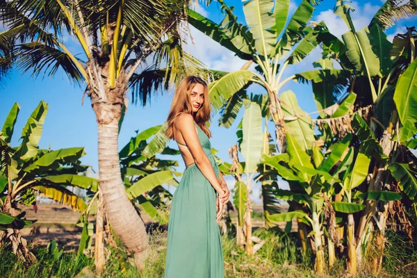 リゾートタウンの緑の熱帯の庭で休んでいる間にカメラを見て長いドレスで自信の若い女性観光客のサイドビュー — ストック写真