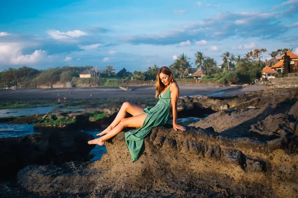 Çıplak ayaklı, ince, güzel yeşil elbiseli bir kadın. Kayalık sahilde oturuyor. Dalgalanan denizin yanında ve başka tarafa bakıyor.