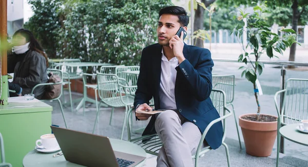Confident hindu male entrepreneur in formal wear talking on mobile phone sitting with laptop computer at terrace, serious businessman working remotely making smartphone call in international roaming