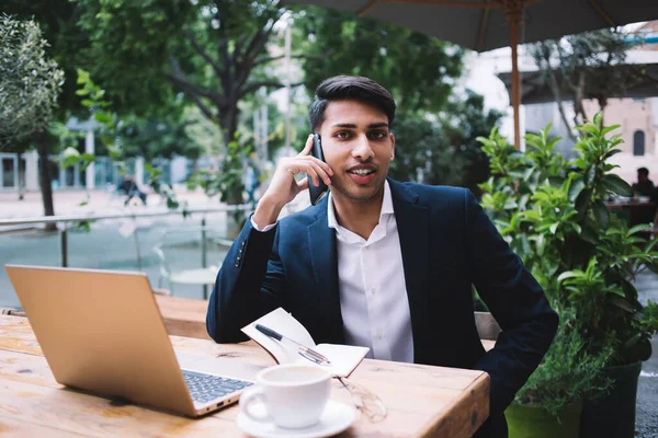 Positive hindu businessman freelancer sitting on terrace making banking via smartphone call in roaming, smiling prosperous male entrepreneur working remotely on laptop computer talking on mobile phone