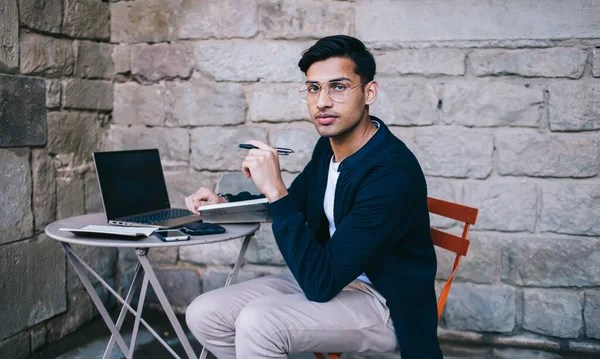 Young casually dressed guy sitting on orange chair in terrace of old building with iron bars on windows and stone walls while working on project