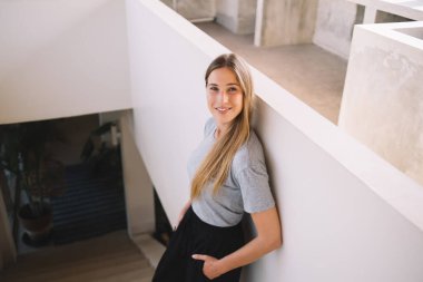 Gri tişörtlü ve siyah pantolonlu güzel bir kadın merdivenlerde elleri ceplerinde duruyor modern iç mekanlarda duvara yaslanıyor ve kameraya gülümsüyor.