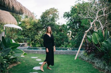 Uzun siyah elbiseli, gülümseyen genç çıplak ayaklı model modern egzotik tatil köyünün yeşil çimlerinde dururken kameraya bakıyor.