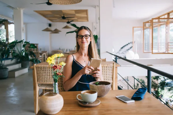 Cheerful adult female in casual clothes and eyeglasses sitting in cafe and smiling with book in hands while eating salad and looking at camera