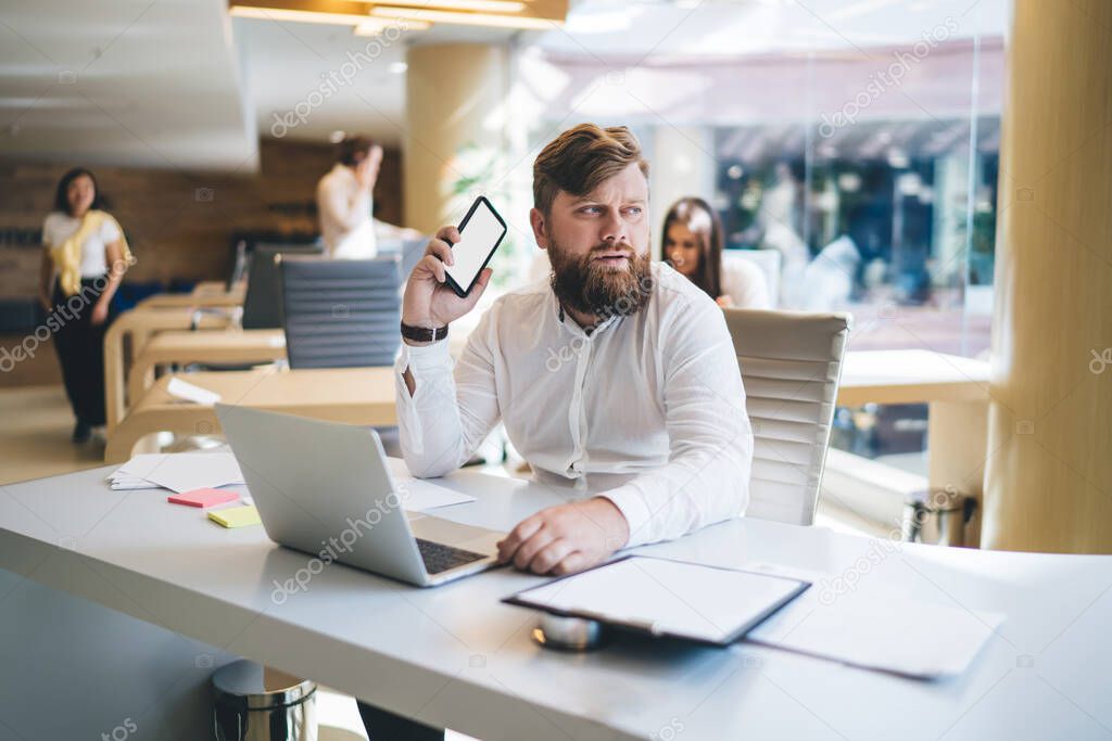 Serious male entrepreneur in elegant wear sitting at table with laptop and using mobile phone while thinking of startup ideas and looking away