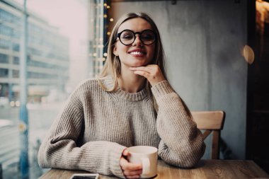 Modern kafenin penceresinin yanındaki masada otururken günlük kıyafetler içinde kahve içen ve kameraya gülümseyen neşeli genç bir kadın.