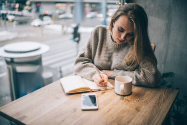 Gündelik giyinmiş zeki bayan öğrenci eğitim kitabına yazı metni yazıyor, kafeterya masasında oturan yaratıcı beyaz kadın organizasyon için not defteri kullanıyor.