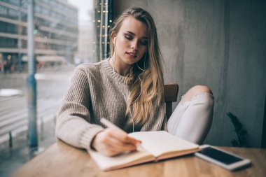 Elektronik kulaklıklı beyaz kadın blogcu eğitim kitabında içerik planı yazıyor. Bilgi verici podcast uygulaması kullanıyor. Kulaklıklı bir kadın kafede kulaklık öğreniyor.