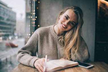 Kulaklıklı olumlu bir genç kadın, başlarını eğip masada oturmuş müzik dinliyor ve modern kafede notlar alıyor.