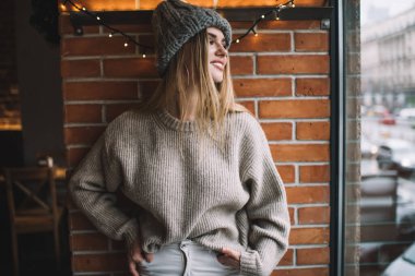 Süveter ve örgü şapka giyen neşeli genç bir kadın. Şehir kafesinde elleri ceplerinde dikilirken gözlerini kaçırıyor.