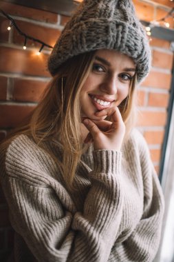 Uzun sarı saçlı, örgü şapkalı, süveterli, gülümseyen ve tuğla duvarın önünde kameraya bakan neşeli genç bir kadın.