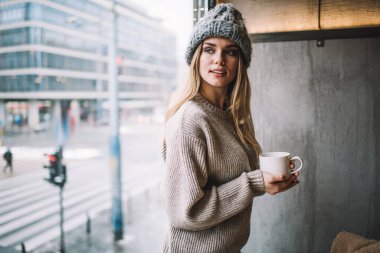 Kokulu kapuçino bardaklı düşünceli hippi kız kafenin içinde kahve molası verirken düşünceli bakışlı, örgü örülü giyinmiş kadın müşteri pencerenin kenarında kafa yoruyor.
