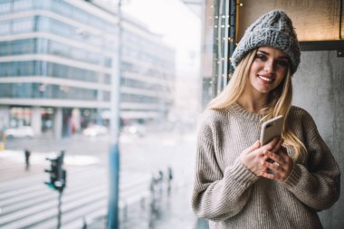 Uzun sarı saçlı, örgü şapkalı, süveterli, gülümseyen ve kameraya bakan genç bir kadın.